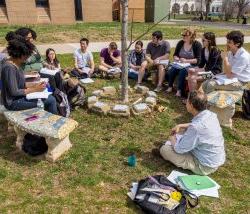 Photo of a classroom in an outdoor setting with faculty and students sitting on benches and the ground.